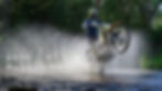 Motorcyclist riding through water, creating a splash. Text reads "2025 Husqvarna Trek, Australia High Country" against a forest backdrop.