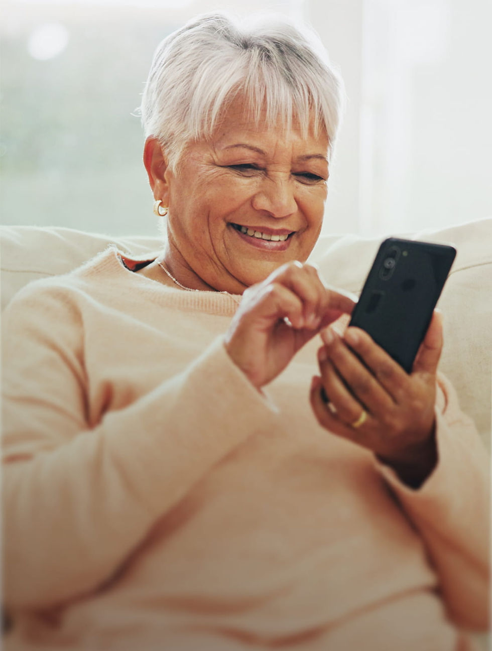 Smiling older woman with short gray hair uses a smartphone, wearing a peach sweater. She's seated, conveying a sense of happiness and relaxation.