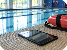 Tablet and rescue tube resting on the deck beside an indoor swimming pool, representing digital near miss reporting and proactive aquatic safety management in a calm, professional facility setting.