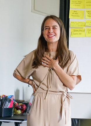 Weibliche Coach, die sich lächelnd ihre linke Hand auf das Herz legt mit Flipchart und Arbeitsutensilien im Hintergrund.