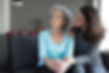 Elderly woman in blue sits on a couch, looking thoughtful. Younger woman comforts her, placing a hand on her shoulder in a cozy living room.
