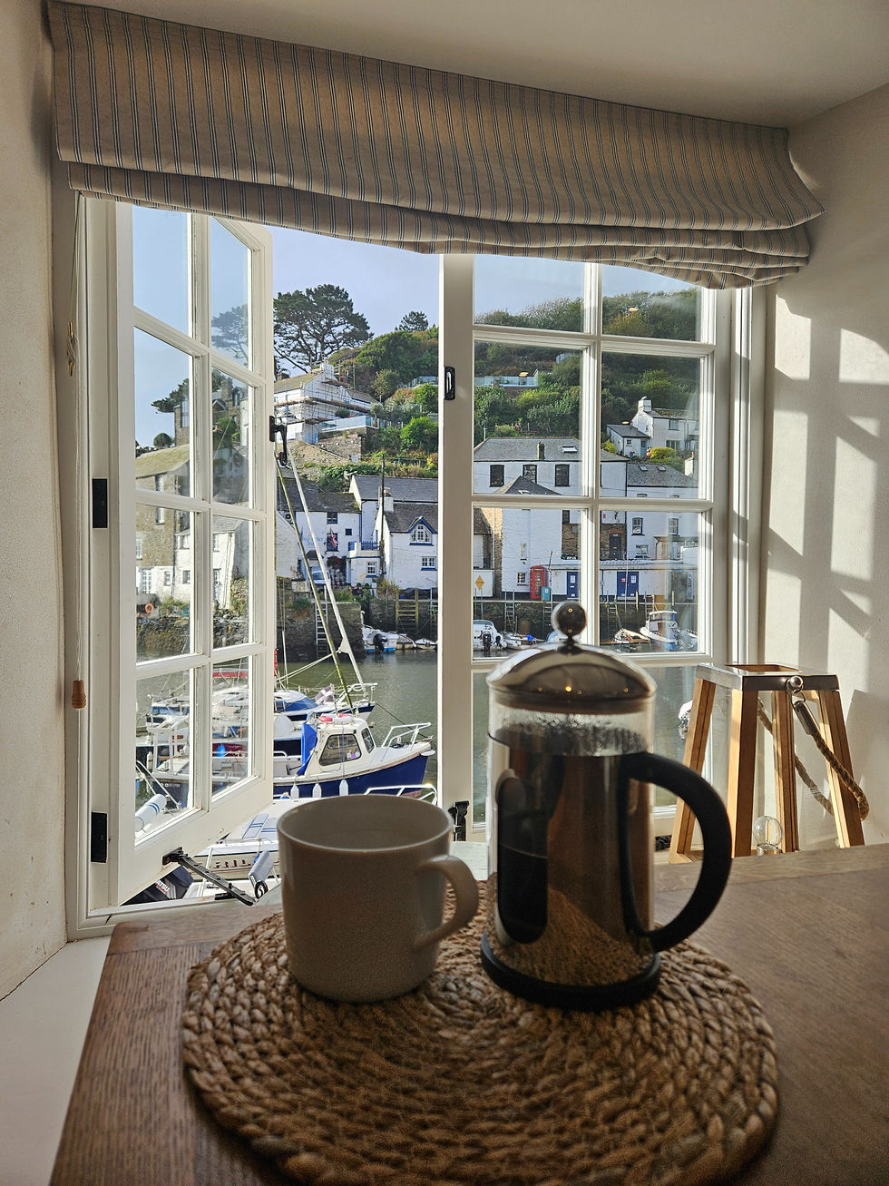 A French press and mug on a table with coastal village view through large window. Calm atmosphere with boats and hillside houses outside.