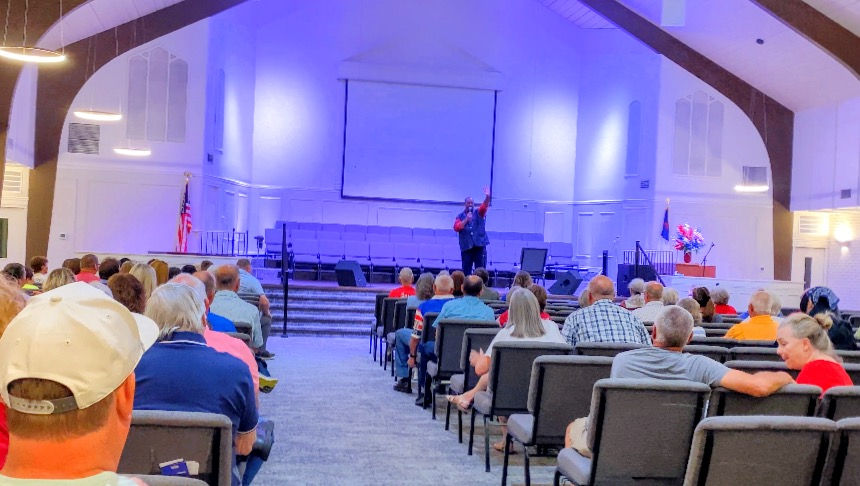 Audience seated in a church auditorium as a speaker stands on a blue-lit stage.