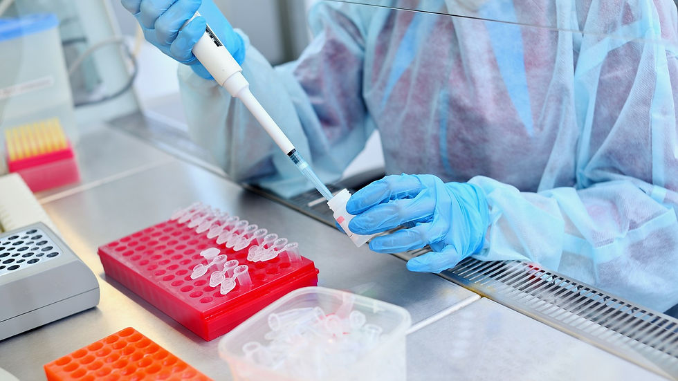 A laboratory technician in a protective gown and gloves pipettes samples into small tubes at a lab bench, with tube racks in the foreground.