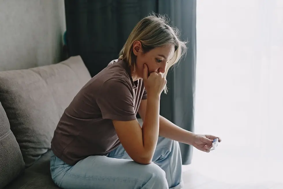 Worried woman sits on a gray sofa, staring at a pregnancy test in a bright home with curtains and soft light.