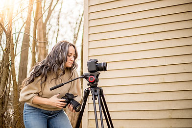 SHE offering her videography services using her cameras and tripod in the forest by a tan building in Medina OH