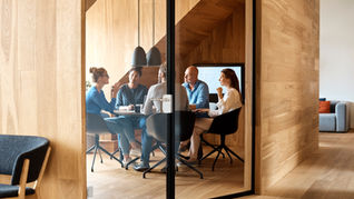 Five people in an office meeting room, engaged in discussion. Wood-paneled walls, modern decor, and a screen in the background.