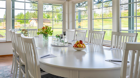 På bilden ser man en del av konferensrummet Birdie på CN Hotell Kallfors. Bilden visar vita möbler. Vid varje plats ligger block och penna och på en bricka står vattenkaraffer och glas. På var sin sida av brickan finns en glasvas med blommor och en fruktskål med äpplen.