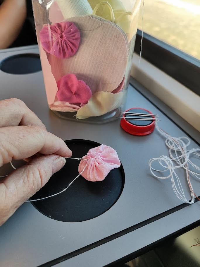 palstic bottle wiht fabric circles and yoyos inside on atable on a train with a hand sewing a pink yoyo