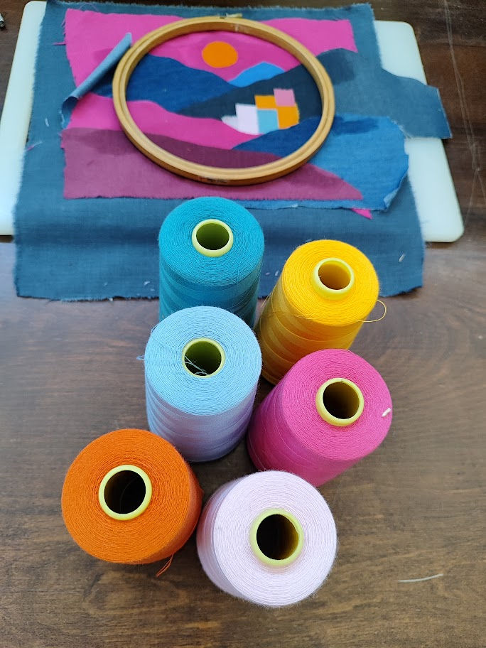 meditteranean summer art quilt pieces iunder a wooden hoop with spools of thread on a table