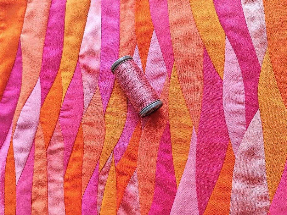 a sunrise colored wave landscape technique mini art quilt with a spool of pink thread lying on top of it