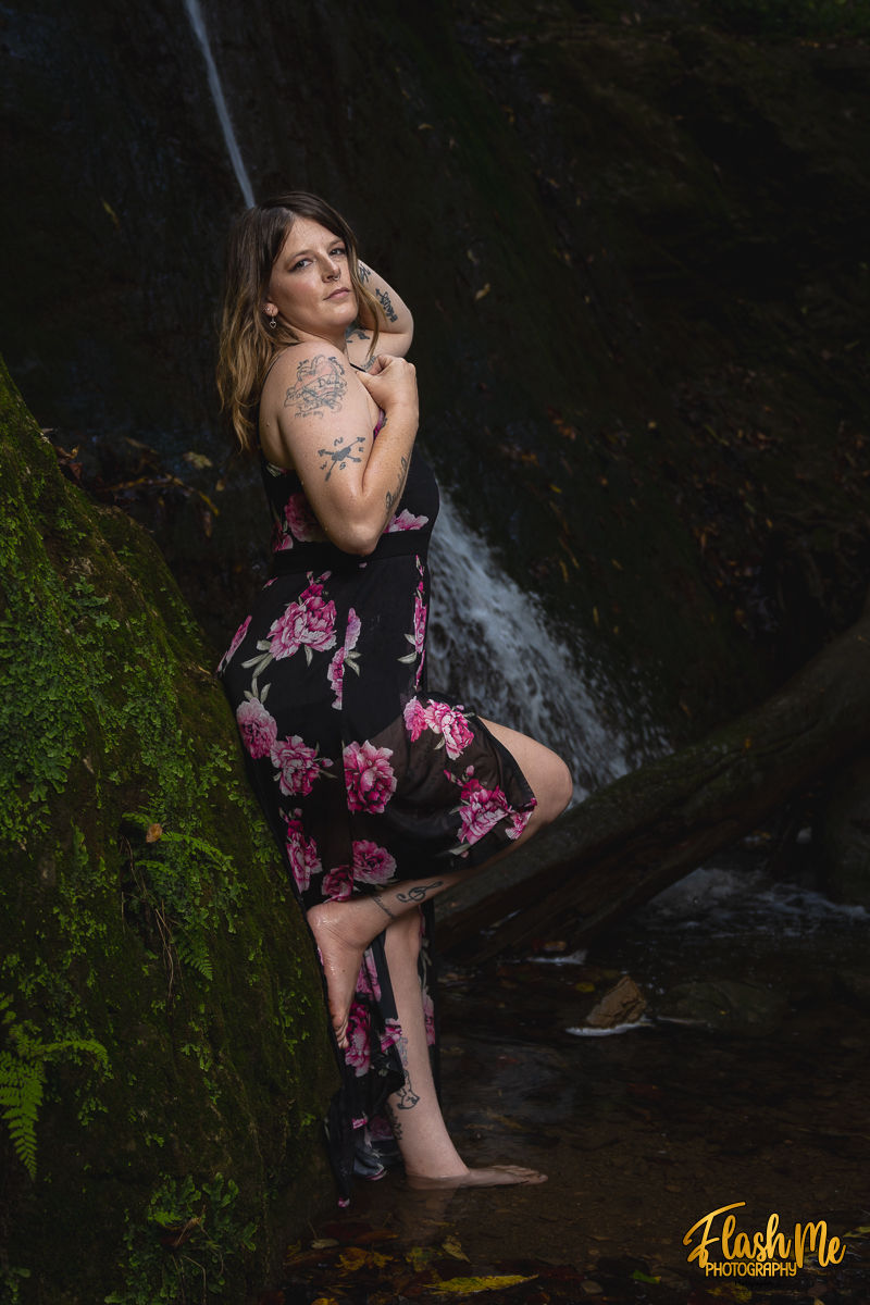 Woman in floral dress poses by a mossy rock and waterfall. Visible tattoos, serene expression. Text: Flash Me Photography.