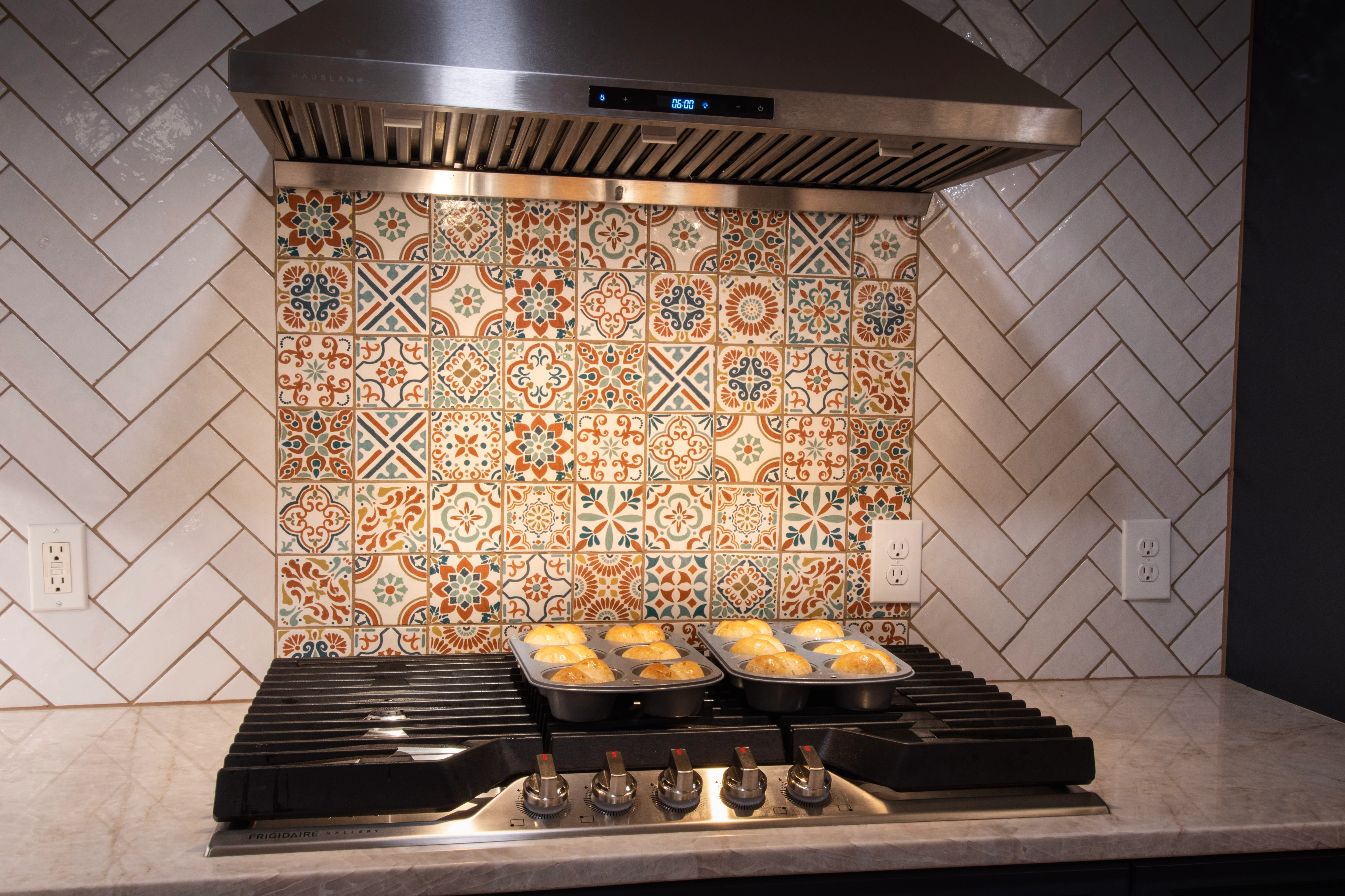 Kitchen with muffins on stovetop, colorful tile backsplash, range hood.