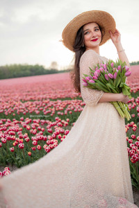 Woman in a hat and long, beige dress holding a bouquet of tulips in tulip field