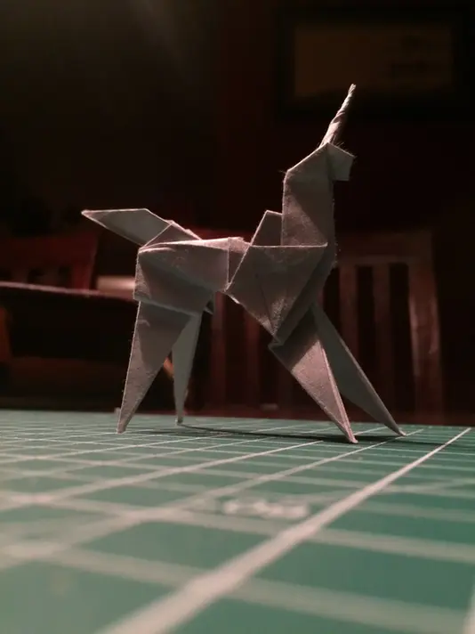 Origami Unicorn on a green grid patterned cutting mat. There is a chair in the backgroun.