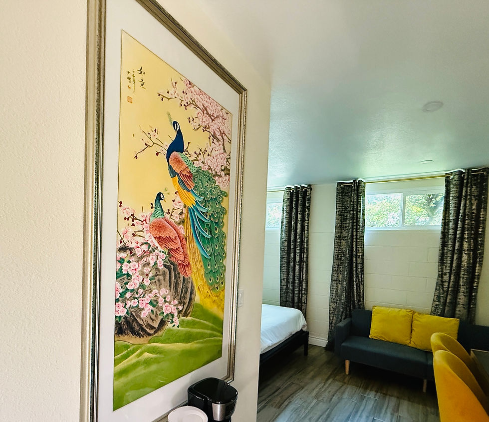 Framed peacock painting with vibrant colors on a wall, next to a cozy room with a bed, gray couch, yellow pillows, and curtained windows.
