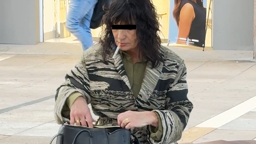 A woman in a patterned jacket sits on a bench, searching her black bag, cigarette in mouth. A poster of a person with glasses in the background.