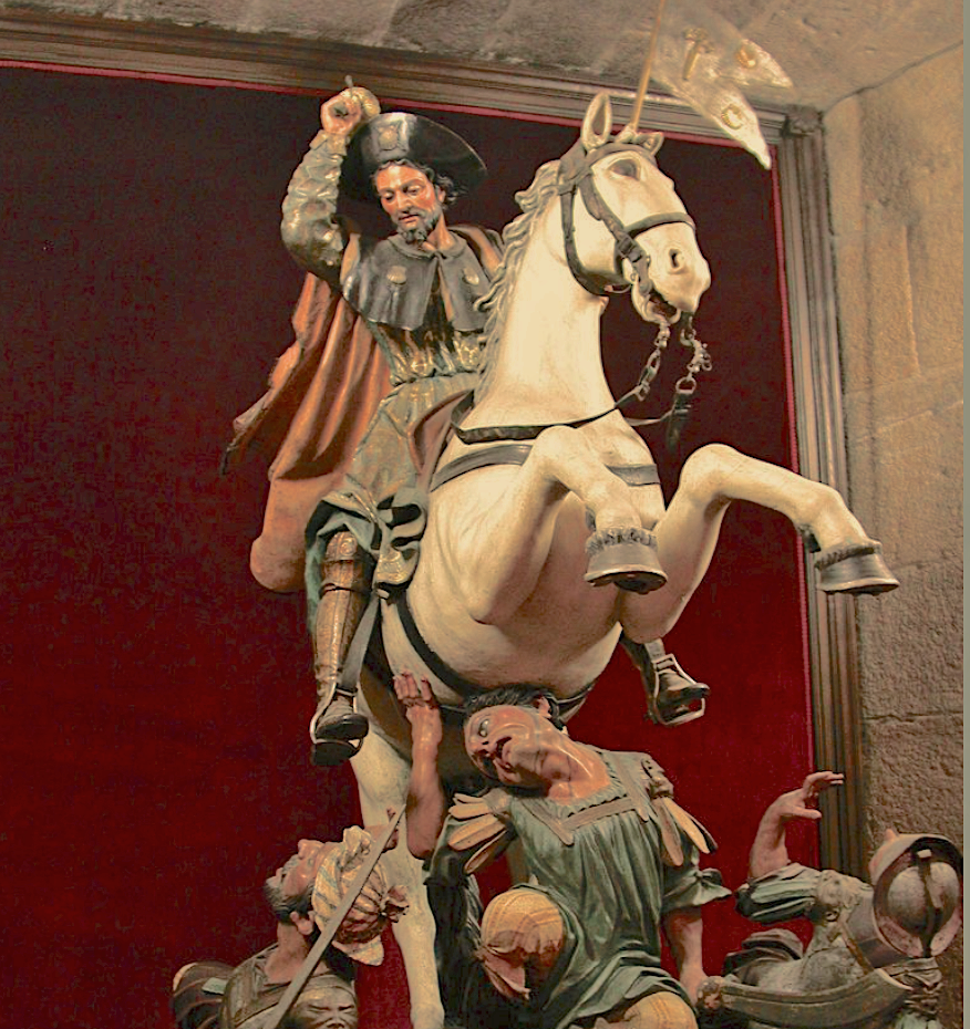 A sculpture of a soldier on a rearing white horse, representing St. James, Matamoros, raising a sword, surrounded by figures on a red and stone background. Dramatic and intense.