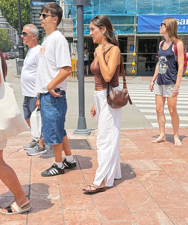 People wait at a sunny city crosswalk, including a man in white and a woman in a brown top and white pants, near blue scaffolding.