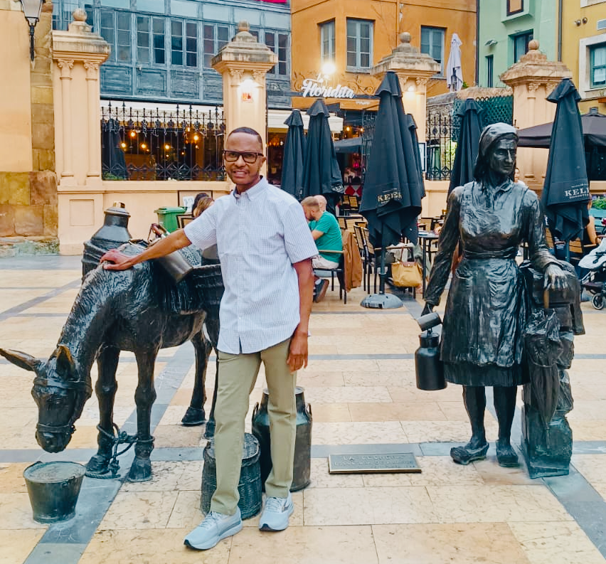 Man smiling next to bronze donkey and woman statues in a plaza in Oviedo, Spain. Cafe in background with black umbrellas. Warm tones and lively atmosphere.