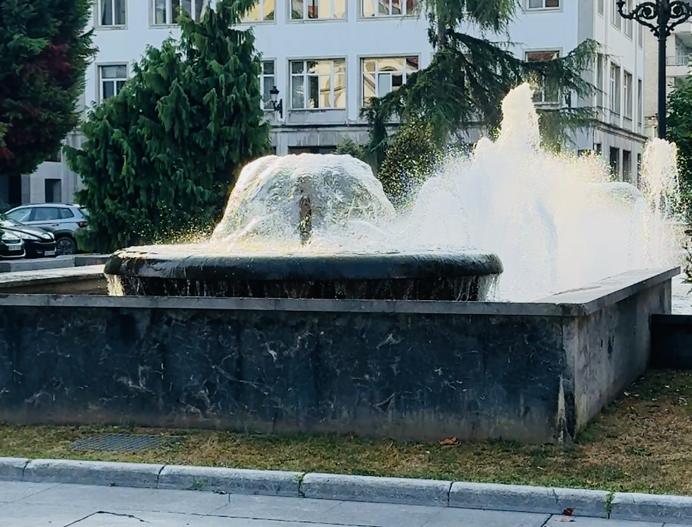 Stone fountain spraying water, surrounded by grass. Background with trees and a building with multiple windows. Bright, sunny day.