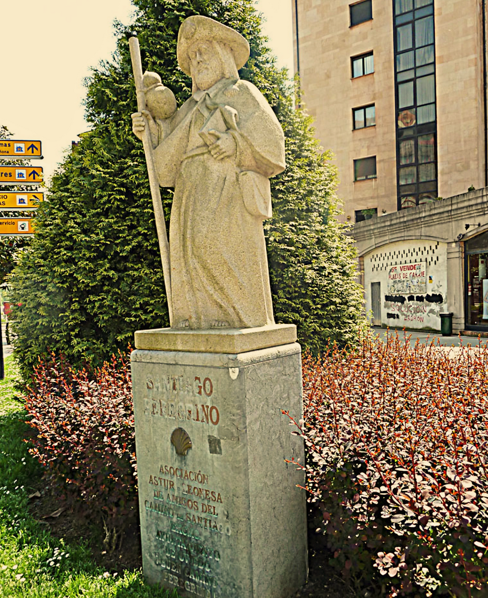 Stone statue of a pilgrim Santiago Peregrino sculpture in La Florida, oviedo, with a staff in a park. Text reads "Santiago Peregrino" on base. Bushes and signs in the urban background.