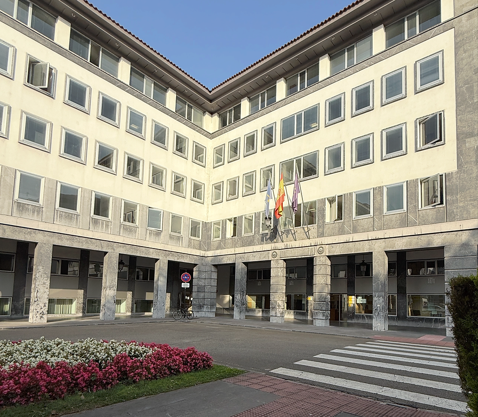 National Police white building, Oviedo, with many windows, flags, and a bike near the entrance. Flower bed and crosswalk in front. Clear sky and calm atmosphere.