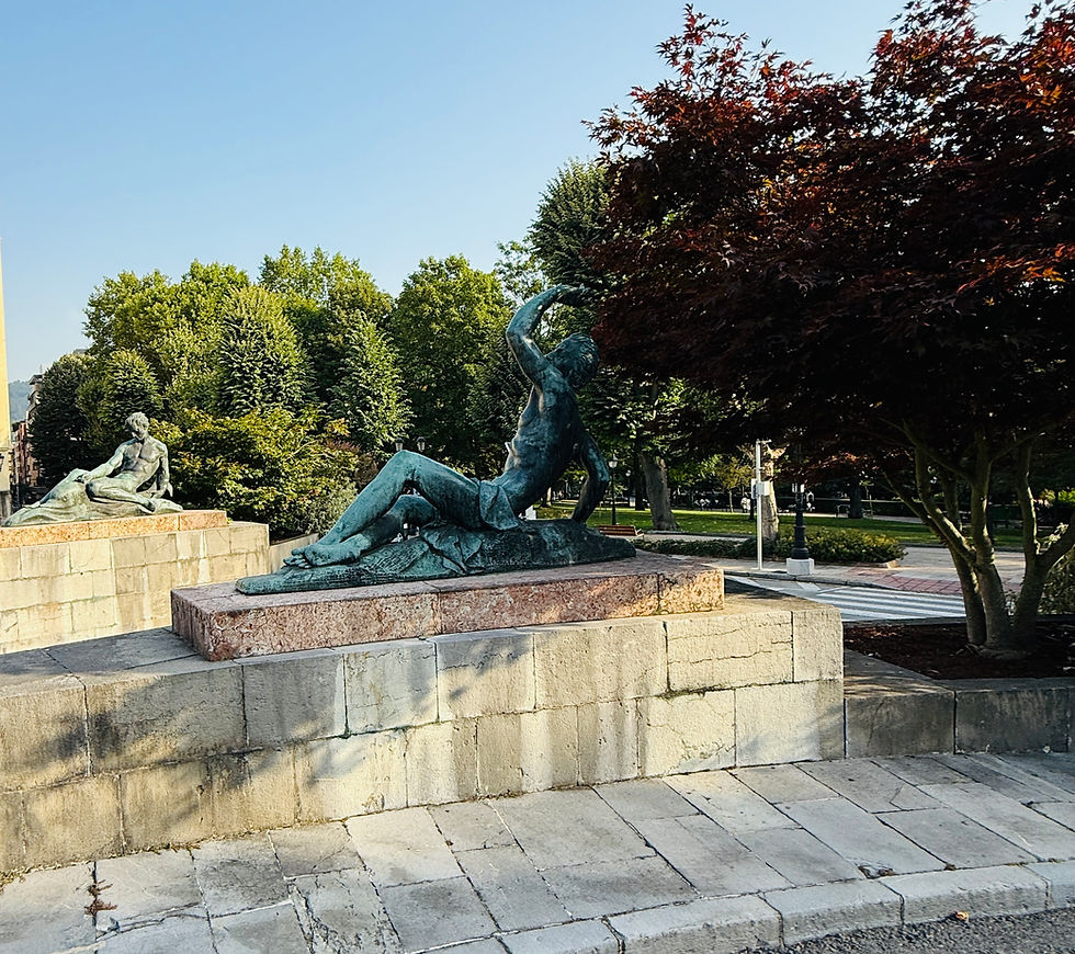 Bronze statues of reclining figures on stone pedestals, surrounded by lush green trees and red foliage under a clear blue sky (Oviedo).
