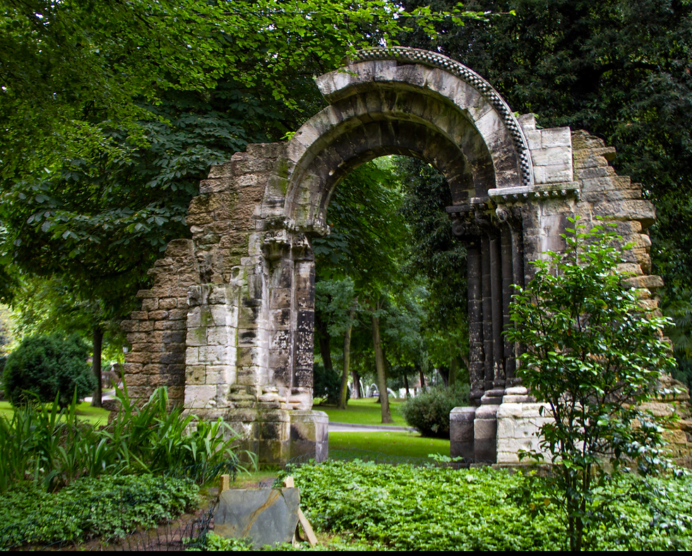 Ancient stone arch ruins surrounded by lush greenery in a park setting. Dense trees and bright green foliage create a serene atmosphere.