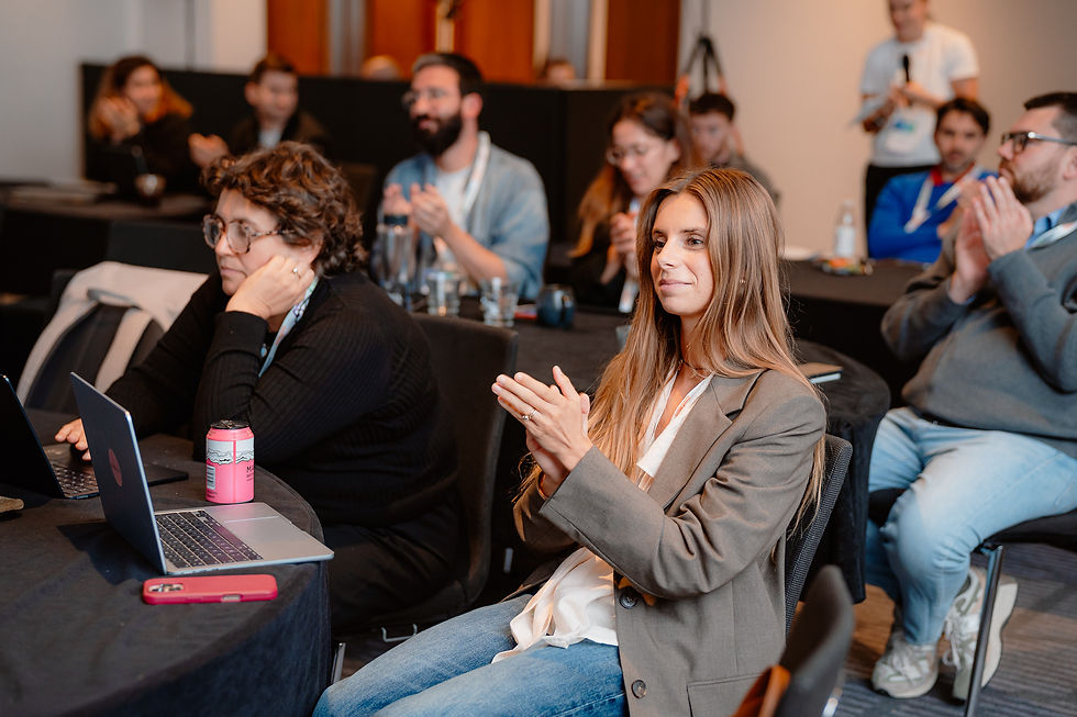 Audience applauds in a conference room, with laptops on tables and a woman in a gray blazer smiling in the foreground.