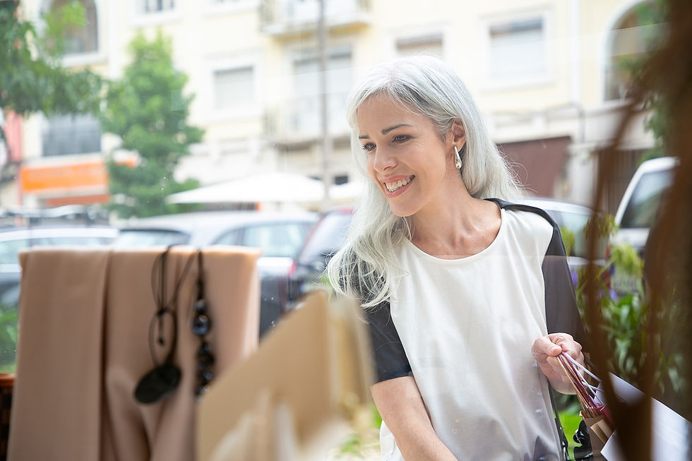 Smiling woman with gray hair window shopping, holding shopping bags. Street view shows cars, trees, and buildings in the background.
