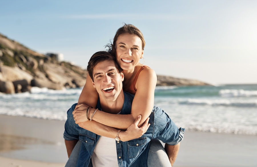 Smiling couple enjoying a piggyback ride on a sunny beach with waves in the background. The mood is joyful and relaxed.