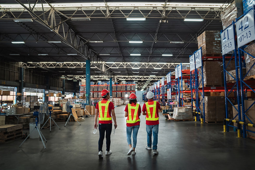 Three workers in red vests and helmets walk in a large, organized warehouse. Rows of shelves with boxes; overhead signs display numbers.