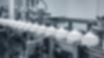 Conveyor with jars of white cream lined up in a factory setting. Metal machinery surrounds the jars, creating an industrial atmosphere.