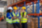Three workers in safety vests and helmets discuss plans in a warehouse. Shelves and boxes fill the background.