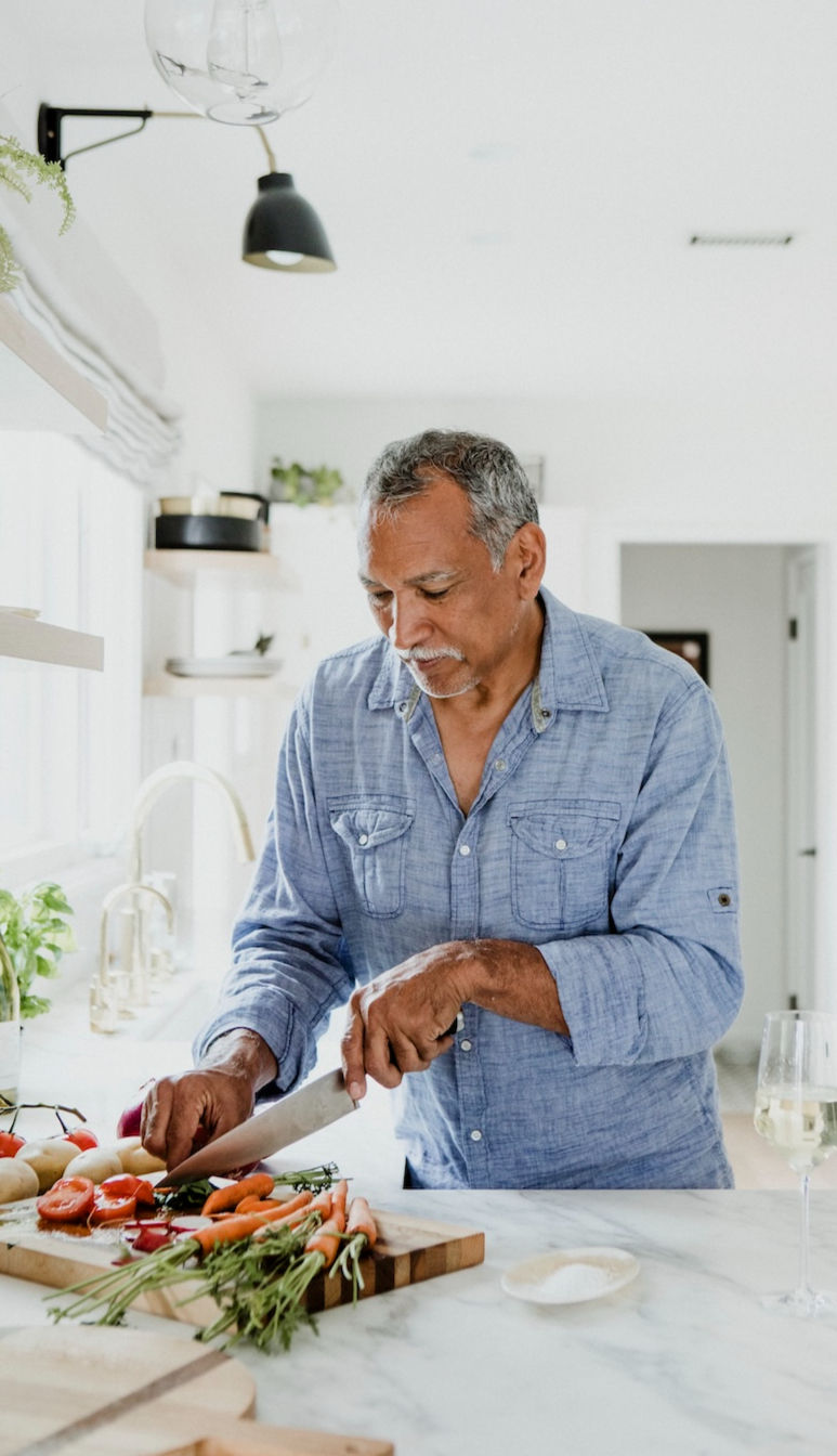 Man in a blue shirt chopping vegetables on a kitchen counter. Bright setting with a glass of white wine nearby, creating a serene mood.