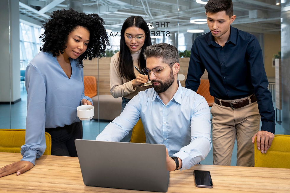 Four colleagues in an office, focused on a laptop. One holds a cup, another takes notes. Bright, modern setting with glass walls.