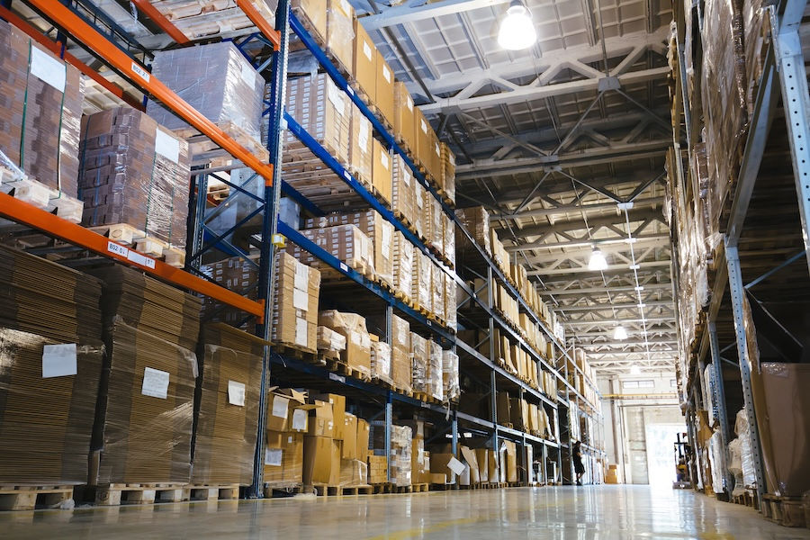 Warehouse interior with tall shelves filled with cardboard boxes and pallets. Overhead lights illuminate the spacious, organized setting.