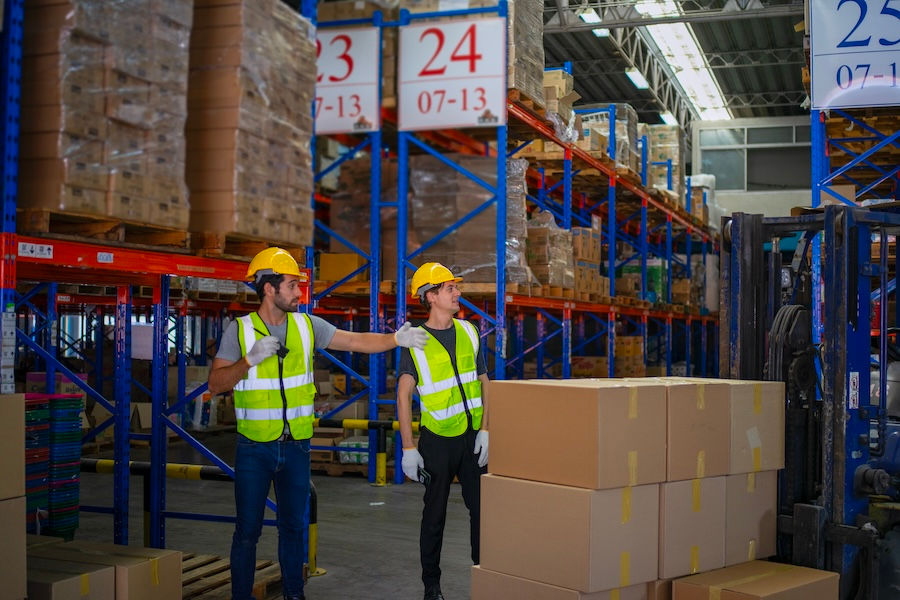 Two warehouse workers in hard hats and safety vests stand by stacked boxes and a forklift under aisle signs 23, 24, 25.