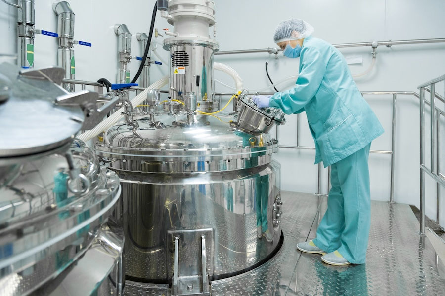 Technician in teal scrubs inspects large metal equipment in a sterile lab. Pipes and machinery fill the bright, industrial setting.