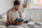 Man reading a book on a bed in a bright room. He wears a beige shirt and jeans. A cactus in a pot is on the windowsill. Cozy ambiance.