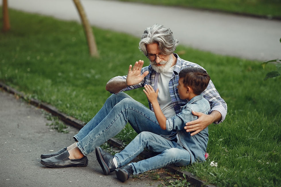 Elderly man and young boy sitting on a park path, smiling and playing hand games. Green grass background, both wearing denim.