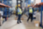 Two workers in yellow vests and helmets organize boxes on shelves in a warehouse. One uses a tablet; the other reaches for a box.