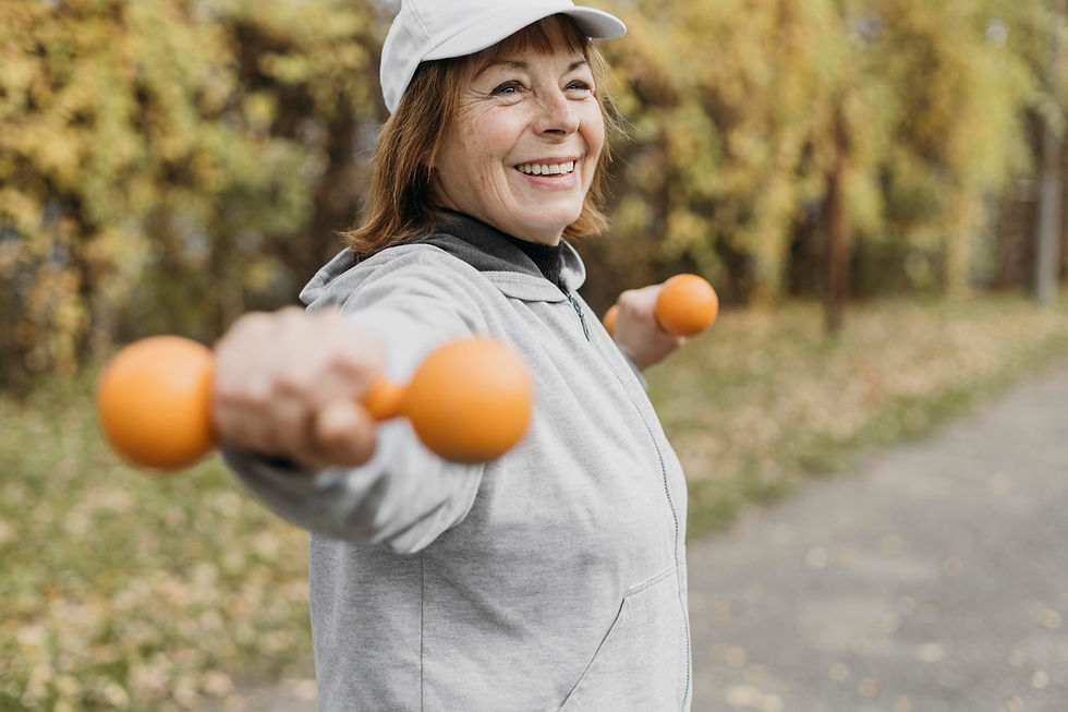 Smiling woman in gray hoodie and cap exercises with orange dumbbells outdoors, leafy background. Energetic and joyful mood.