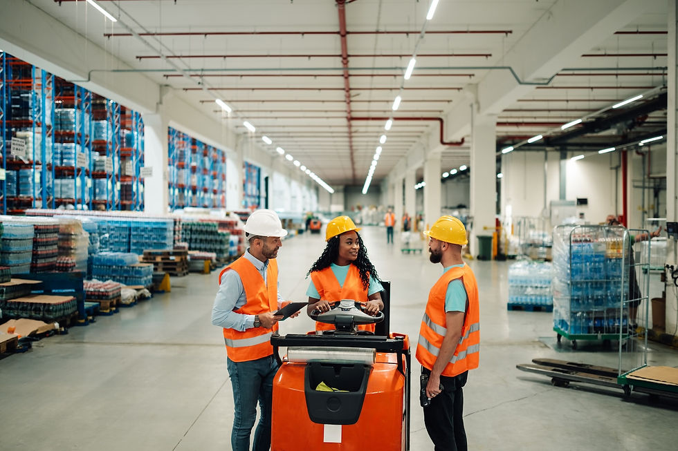 Three workers in orange vests, chatting by a forklift in a bright warehouse. Shelves with products line the walls, creating a busy scene.