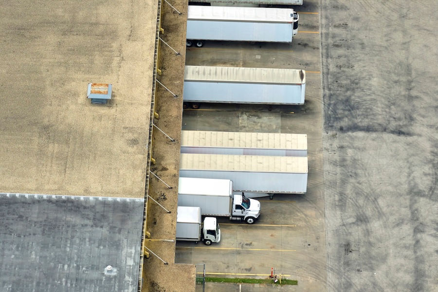 Aerial view of multiple freight trailers and delivery trucks parked at loading docks beside a large warehouse.