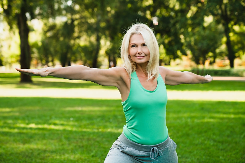 Woman in a green tank top doing yoga with outstretched arms in a sunny park, surrounded by green grass and trees, looking calm.