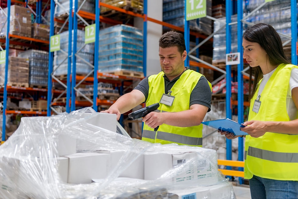 Two workers in bright vests scan boxes in a warehouse aisle. Shelves are stocked with goods. They appear focused and are using a tablet.