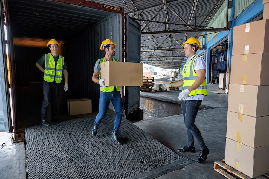 Workers in safety gear unload boxes from a shipping container in a warehouse. One man carries a box, another stands inside, smiling.
