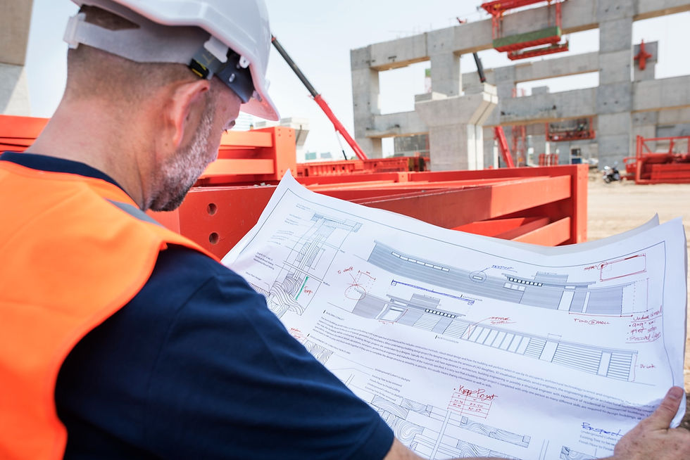 Worker in a hard hat and orange vest reviews blueprints at a construction site. Red scaffolding and concrete structures in the background.